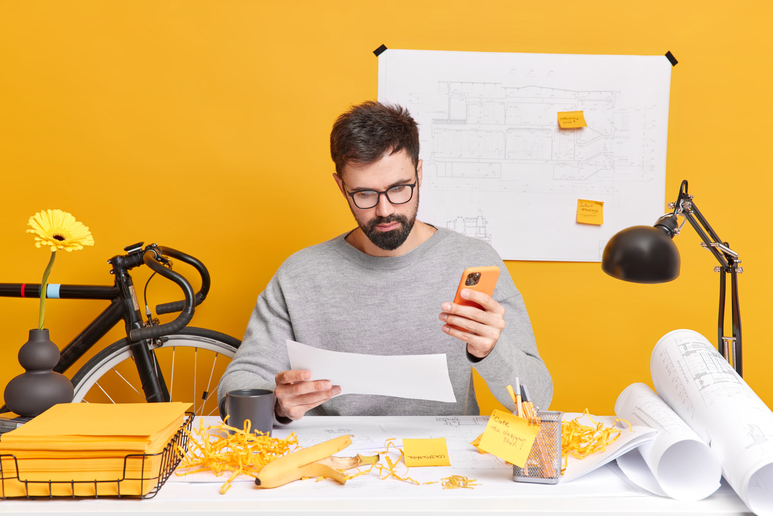 Serious bearded European man concentrated at paper uses mobile phone checks newsfeed makes drawings for construction company surrounded with blueprint rolls stickers and papers. Job concept.