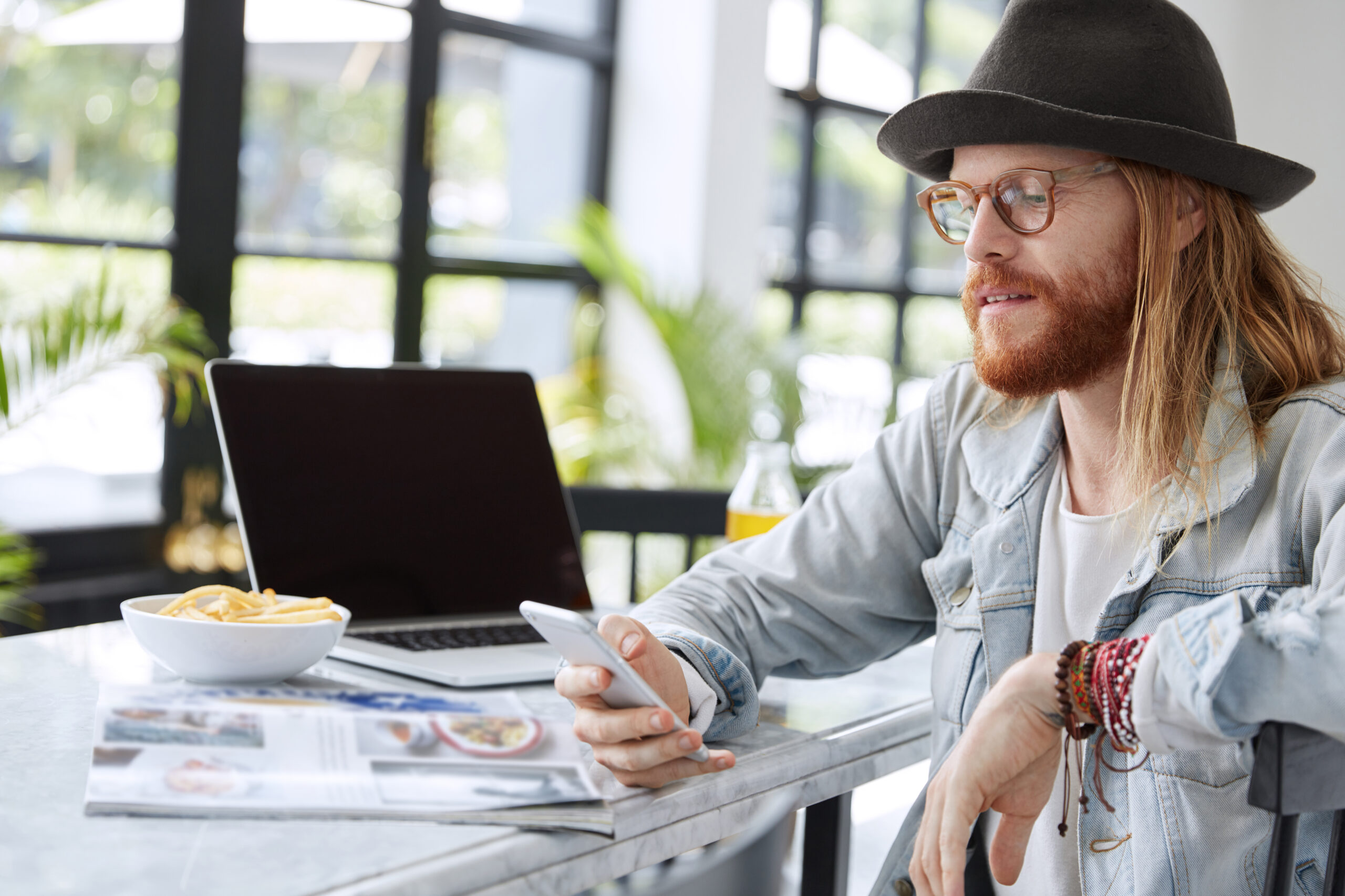 Bearded hipster guy in eyewear and hat, reads text message on smart phone, sits in spacious room, reads magazines, uses laptop computer. Fashionable copywriter checks work of new application